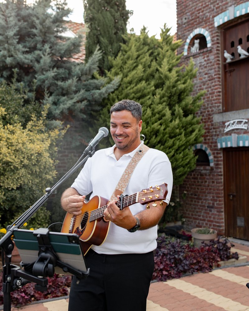 Kevin Hellot souriant, au chant et à la guitare devant une bâtisse en briques